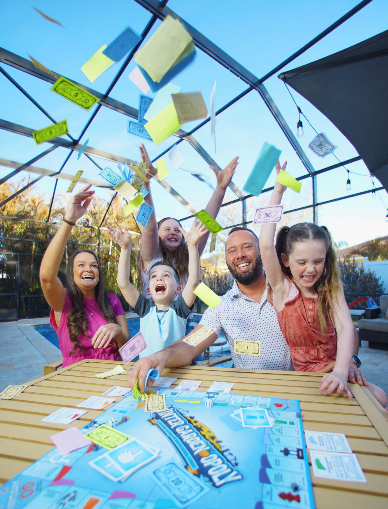 Austin, Kellie, and their three children throwing Monopoly money into the air around their patio table