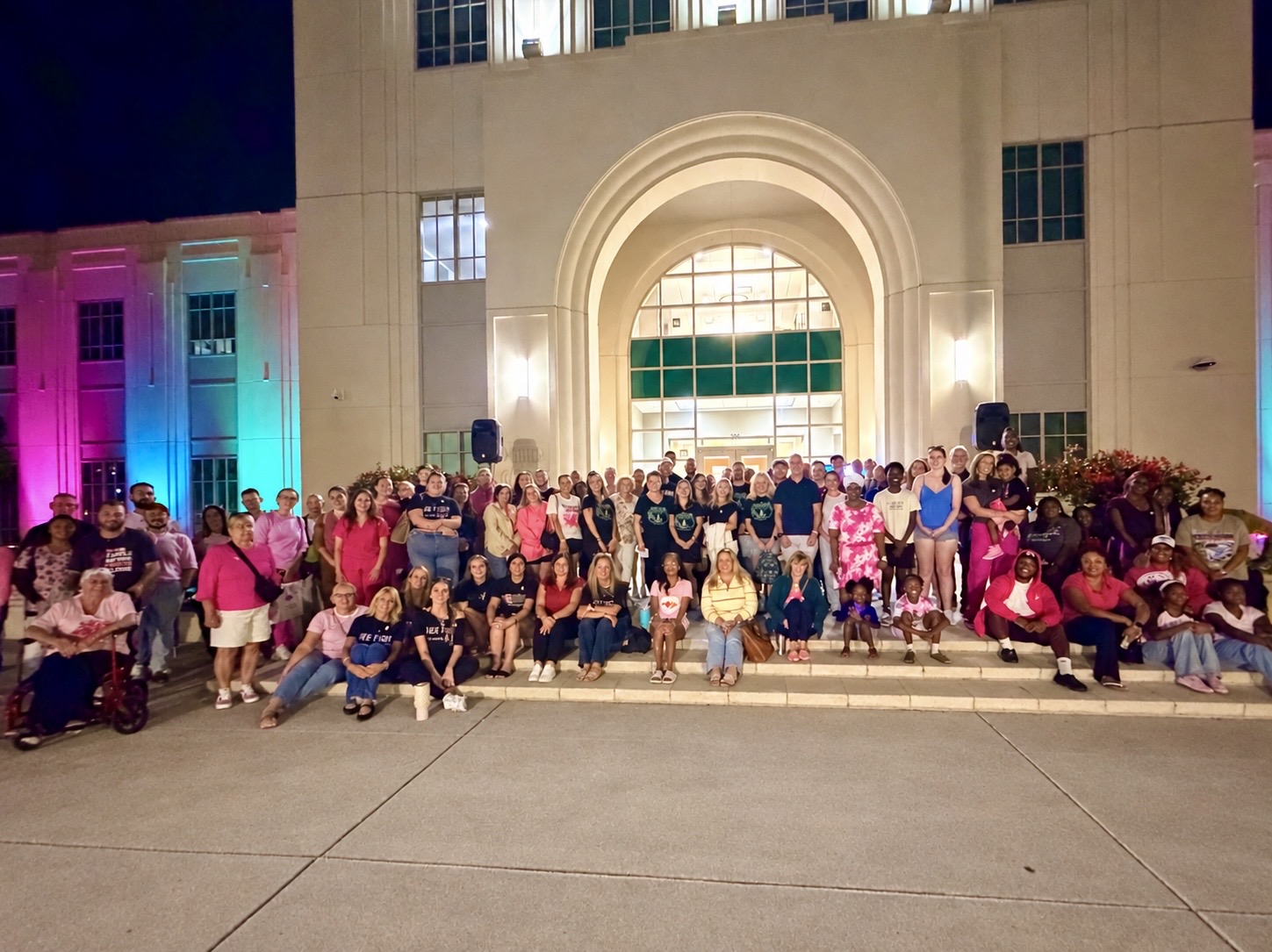 Community members gather on the steps of a Winter Garden building lit in pink and teal for a metastatic breast cancer awareness event